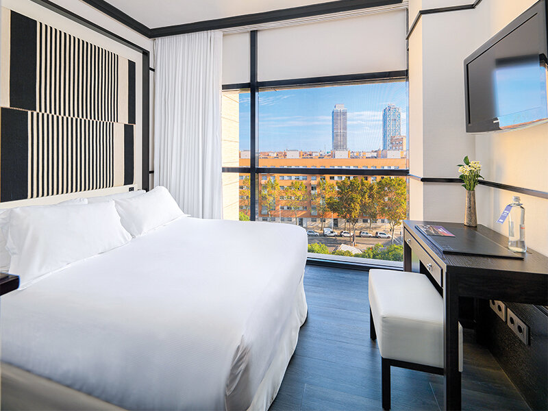 Habitación de hotel moderna con cama blanca, escritorio y gran ventana con vistas.