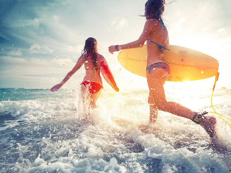 Dos mujeres en bikini con tablas de surf corren por las olas salpicadas hacia el amanecer.
