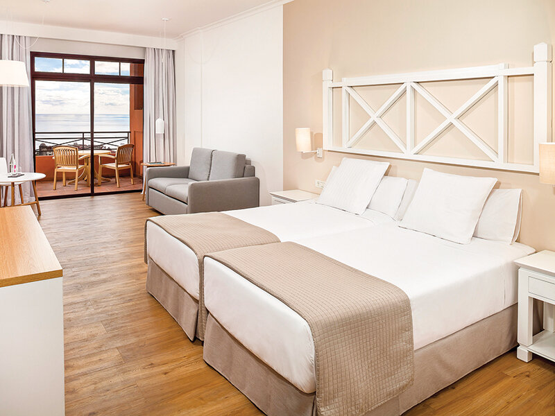 Una habitación de hotel luminosa con cama doble, sofá gris, suelo de madera y balcón con vistas al mar.