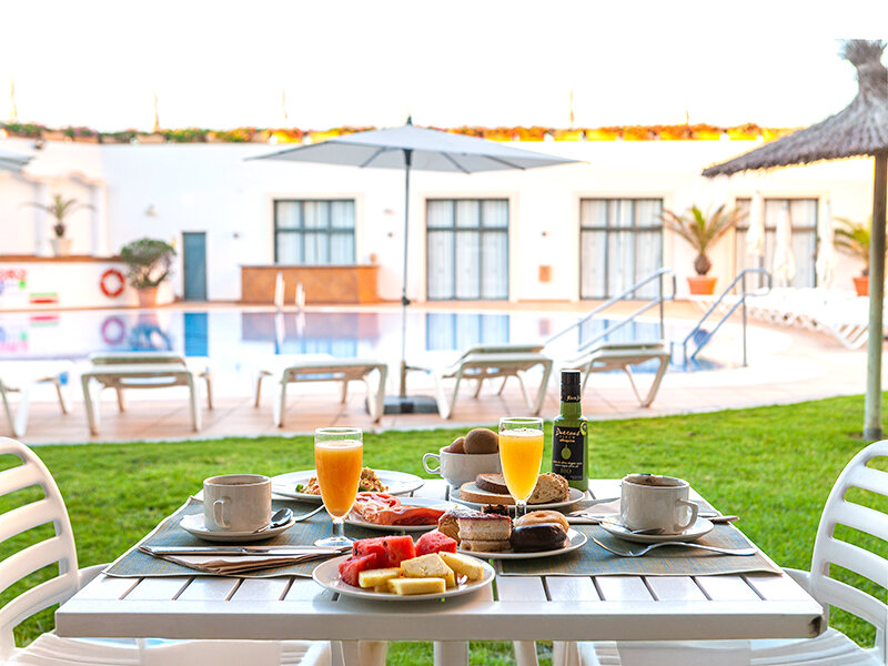 Un desayuno abundante en una mesa al aire libre con vista a una piscina y sombrillas.