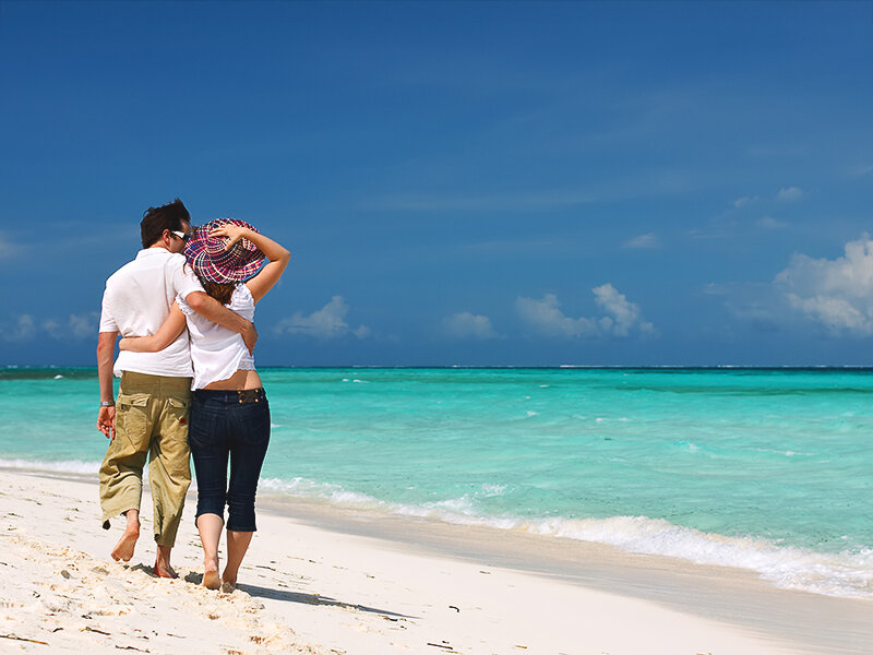 Pareja con ropa blanca caminando por una playa de arena con mar azul y cielo.