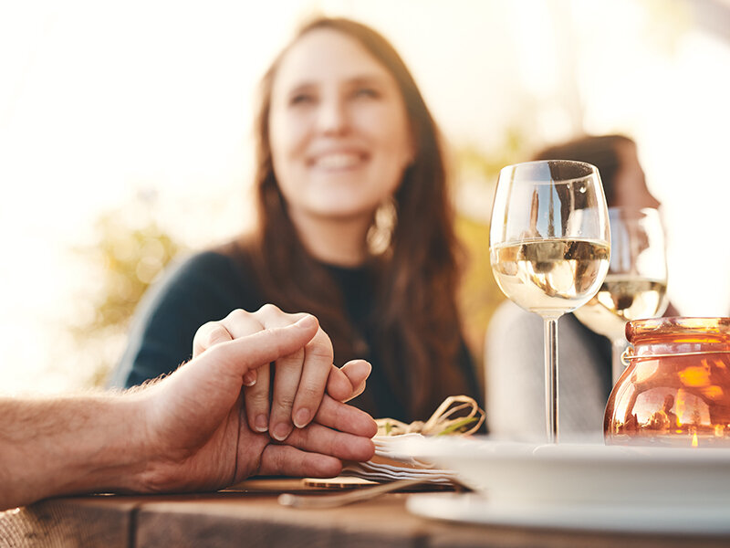 Una pareja se coge de la mano durante la cena, con una copa de vino y una vela en primer plano.