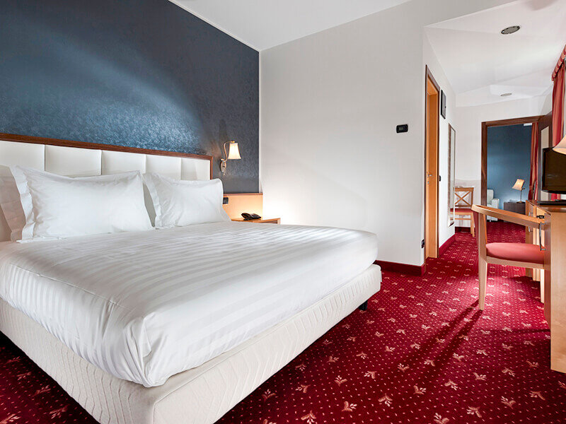 Una habitación de hotel con cama grande, sábanas blancas, alfombra roja estampada y vista a una habitación contigua.