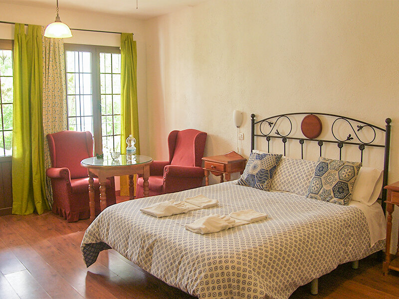 Acogedor dormitorio con cama grande, sillones rojos, mesita y ventanales con cortinas verdes.