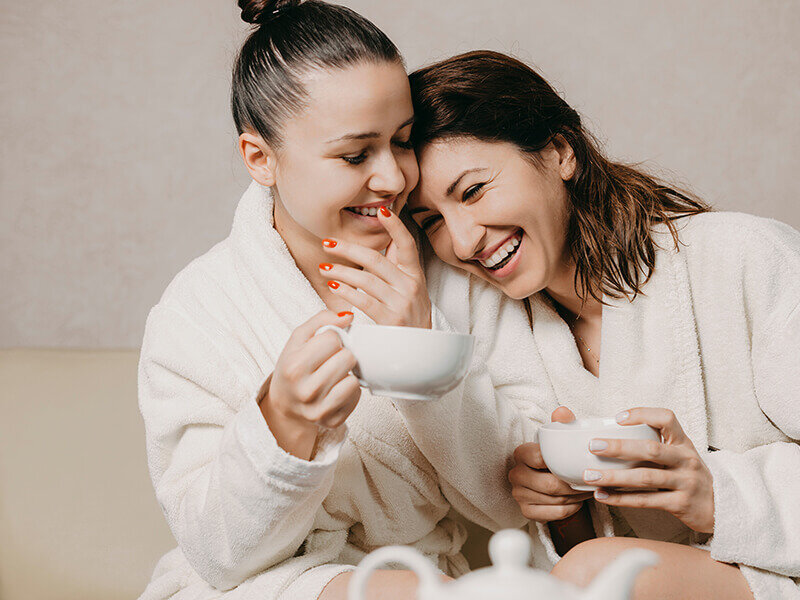 Dos mujeres sonrientes con batas blancas sostienen tazas de café y se sientan juntas.