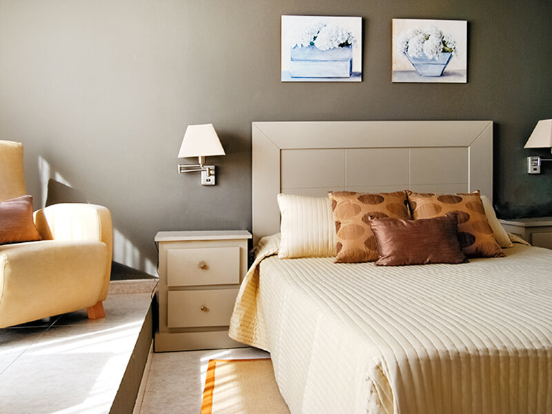 Acogedor dormitorio con cama grande, mesita de noche, apliques de pared y sillón amarillo.