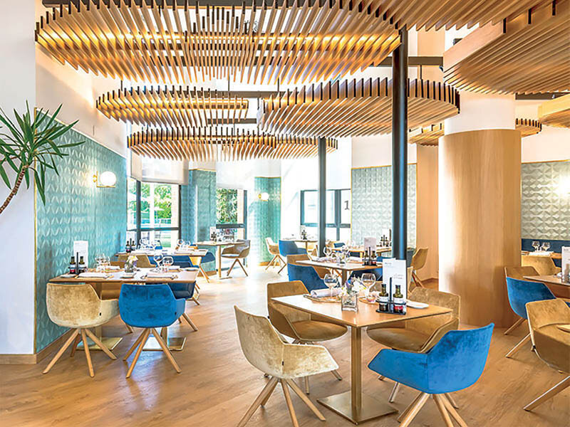 Interior de restaurante moderno con mesas de madera, sillas azules y beiges, y techos de listones de madera.