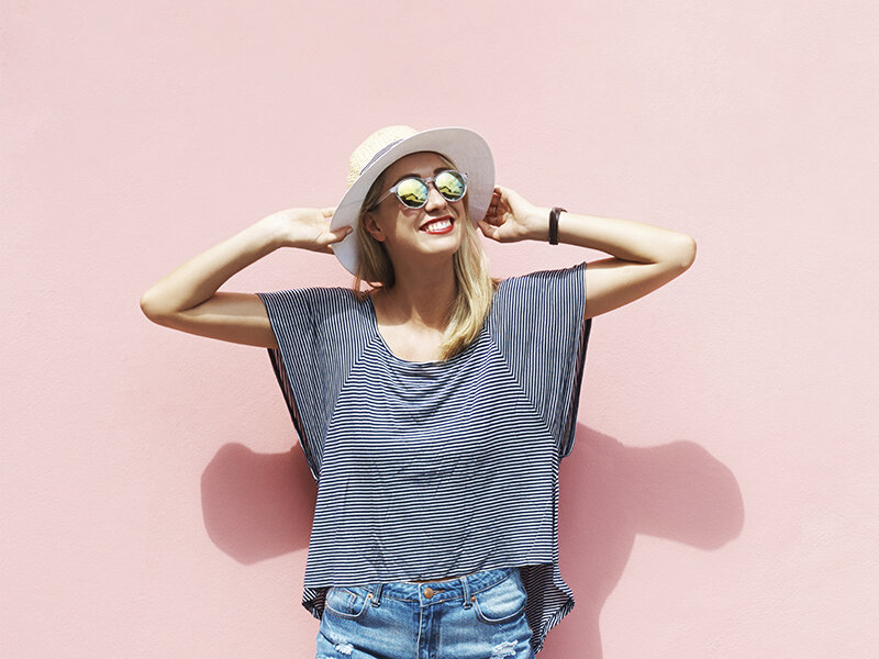 Una mujer rubia sonriente con gafas de sol y sombrero posa ante una pared rosa.