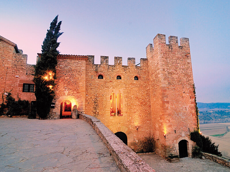 Un castillo de piedra con torres almenadas se ilumina al atardecer, con un patio empedrado y un ciprés.