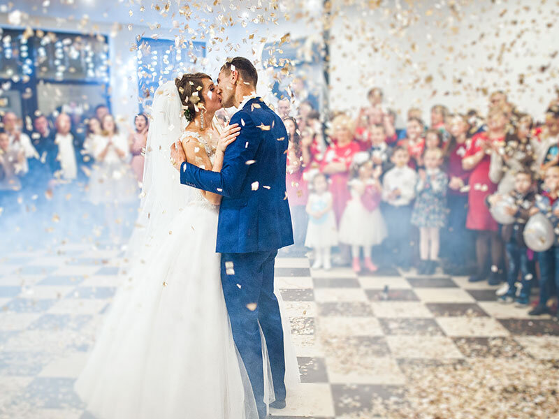 Una pareja de novios se besa en una celebración mientras caen confeti dorado y humo.