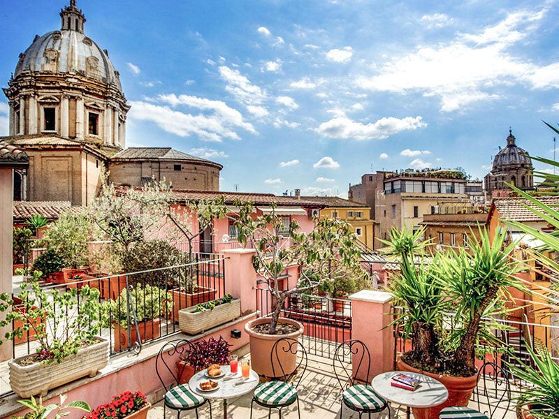Soleada terraza en la azotea de Roma con plantas verdes, mesas pequeñas y un gran edificio con cúpula.