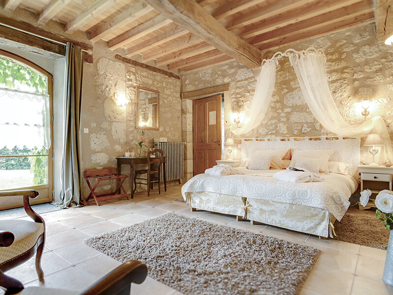 Dormitorio rústico con paredes de piedra, vigas de madera, cama grande con dosel y puerta de cristal.