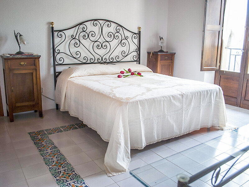 Dormitorio con cama de hierro forjado, ropa blanca, mesitas de noche de madera y suelo con cenefa de azulejos.
