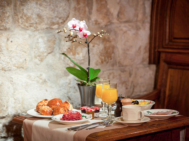 Mesa de desayuno con zumo de naranja, bollería, embutidos, café y una orquídea blanca frente a una pared de piedra.