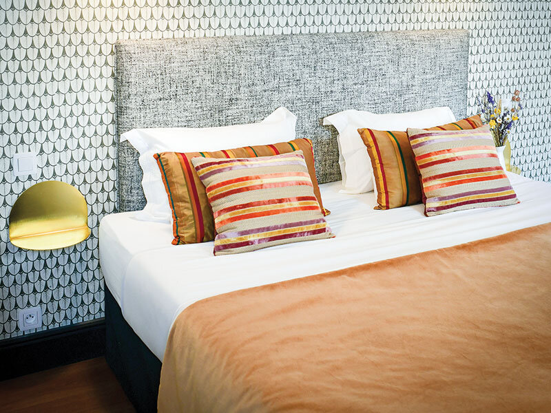 Una cama con ropa blanca, cojines coloridos, una manta naranja y papel pintado con patrones en la pared.