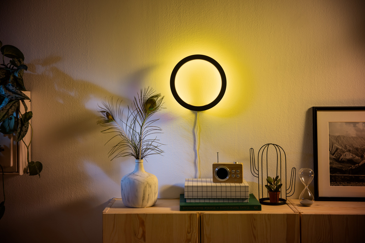 Eine runde Wandlampe leuchtet gelb über einem Holzschrank mit Vase, Radio, Pflanze und einem gerahmten Bild.