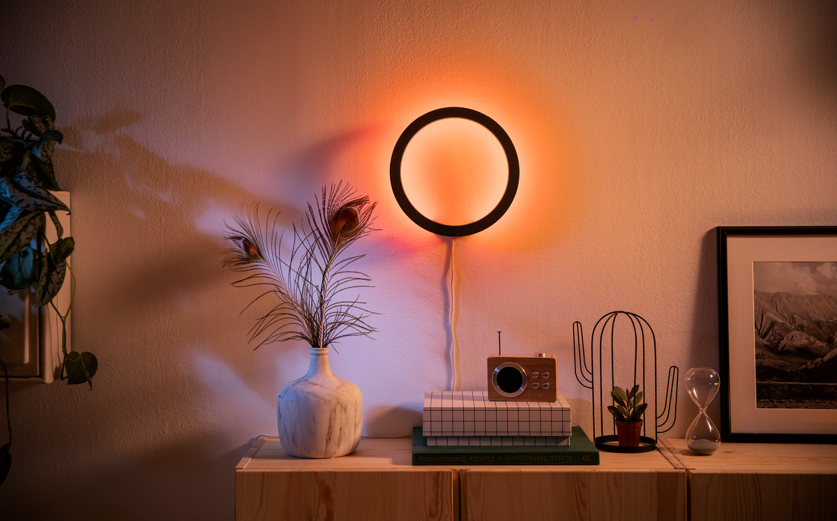Eine runde Wandlampe leuchtet orange über einem Holzschrank mit Vase, Radio, Kaktus und einem Bild.