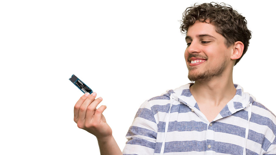 A smiling young man with curly hair wearing a blue and white striped shirt holds a small SSD in his hand.