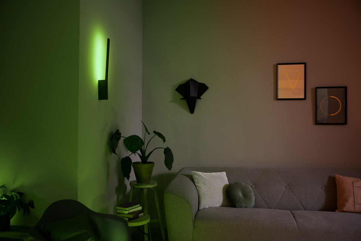 Wohnzimmer mit grüner Akzentbeleuchtung, grauem Sofa mit Kissen, Topfpflanze auf Hocker und Wandbildern.