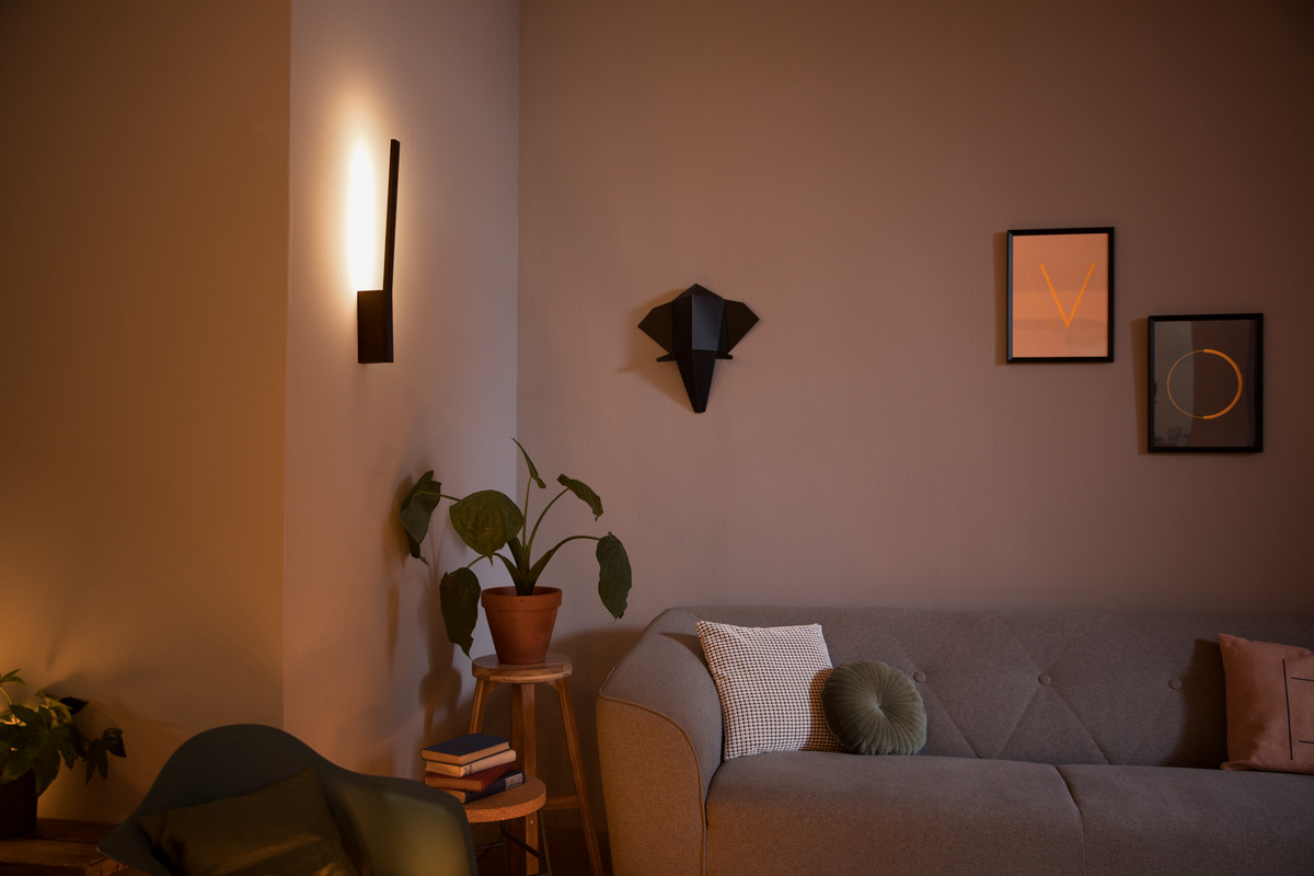 Ein warm beleuchtetes Wohnzimmer zeigt ein graues Sofa, eine Wandlampe, Bilder und Topfpflanzen.