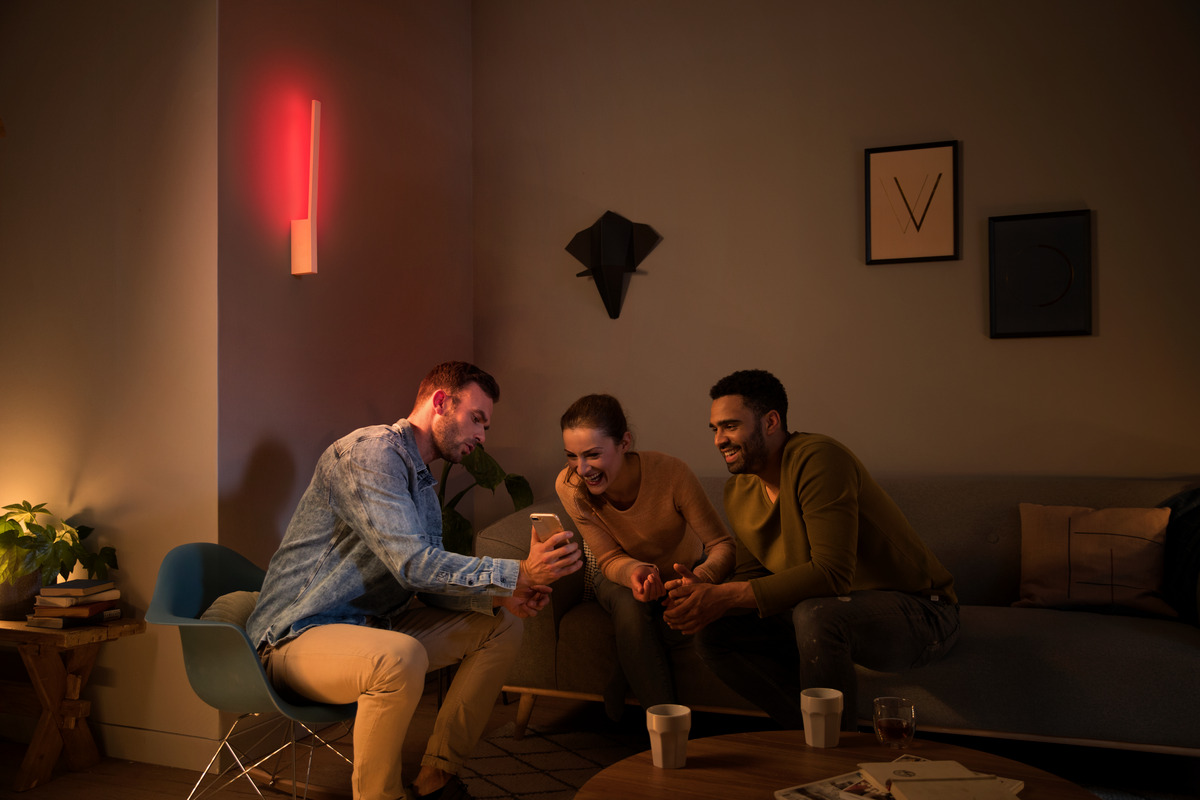 Drei Freunde sitzen im gedimmten Wohnzimmer, ein Mann zeigt etwas auf seinem Smartphone, ein rotes Wandlicht leuchtet auf.