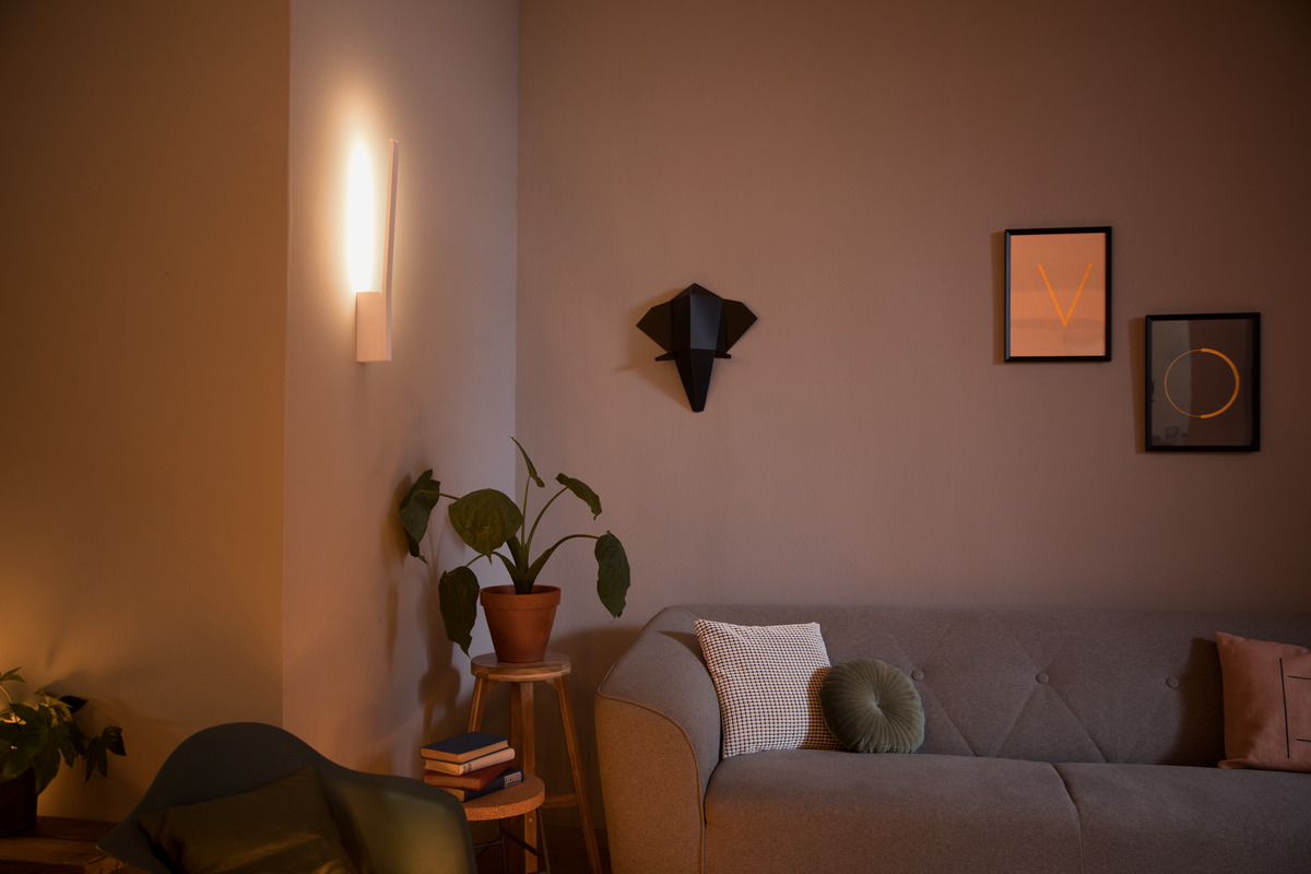 Warm beleuchtetes Wohnzimmer mit grauem Sofa, Wandkunst, Pflanze auf einem Hocker und Wandlampe.