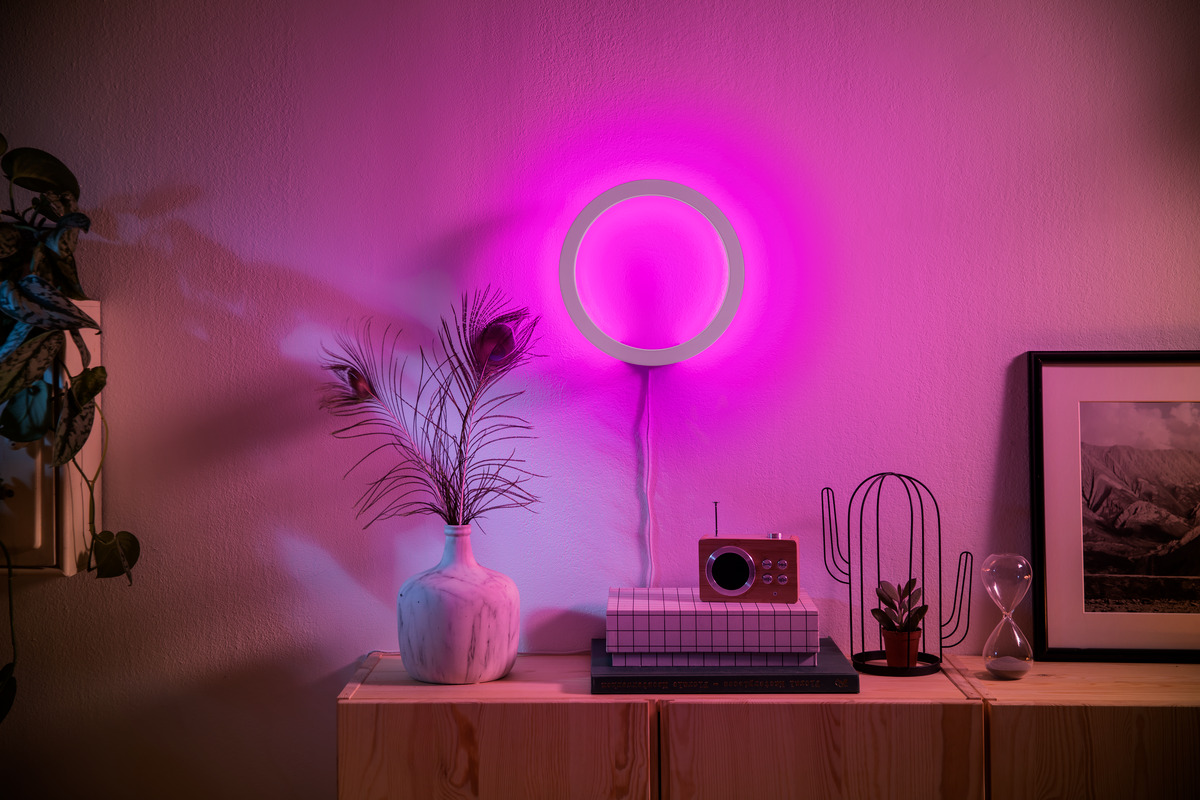 Wohnzimmer mit runder rosa LED-Wandleuchte, Vasen mit Pfauenfedern und Deko auf Holzkommode.