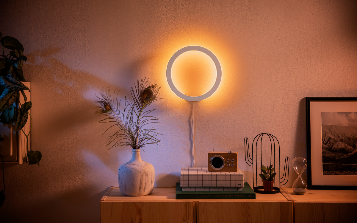 Eine runde Wandleuchte strahlt warmes Licht aus, darunter stehen eine Vase mit Pfauenfedern und ein Radio.