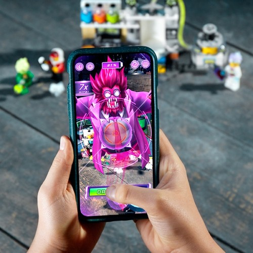 Ein Smartphone in Händen zeigt ein AR-Spiel mit einem rosafarbenen Monster, vor Lego-Sets auf Holz.