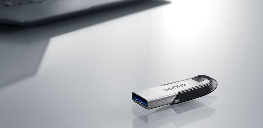 Srebrno-czarna pamięć USB SanDisk spoczywa na błyszczącej szarej powierzchni, z rozmytym laptopem w tle.