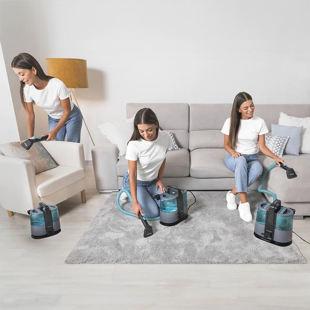 Una donna con maglietta bianca e jeans usa un aspiratore per pulire poltrona, tappeto e divano.