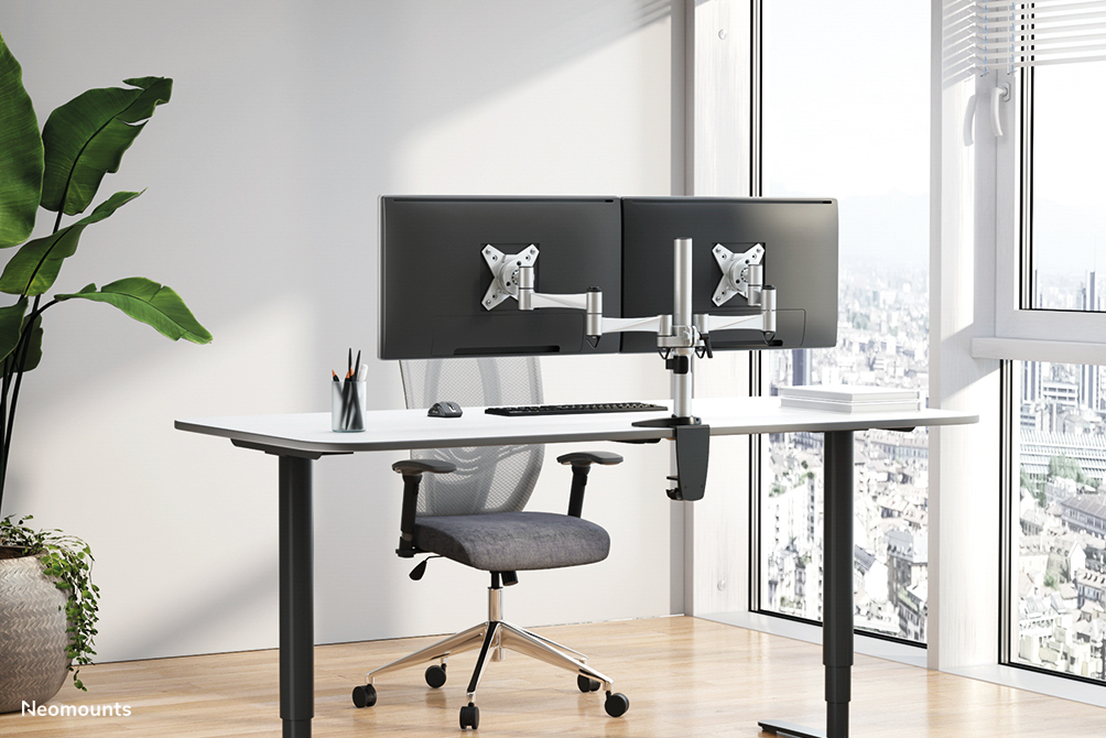 Two monitors on a silver desk mount, on a white desk with a chair, in front of a large window.