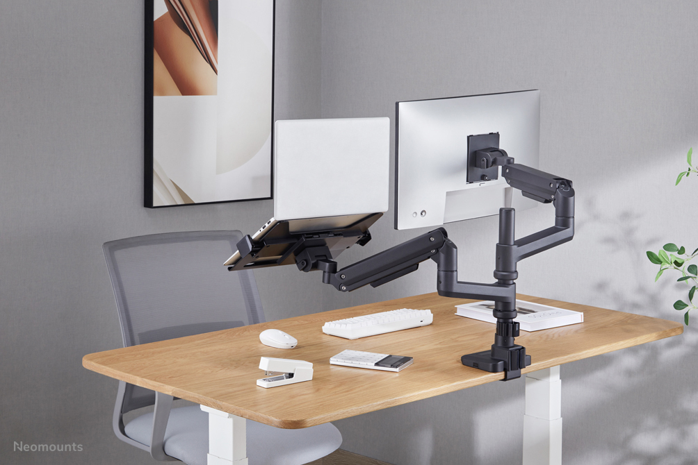 An ergonomic dual monitor arm with a laptop and screen on a wooden desk in a modern office.