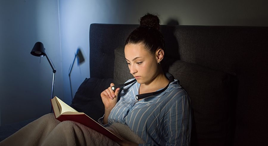 Eine Frau im Bett liest ein Buch mit einer Nackenlampe.