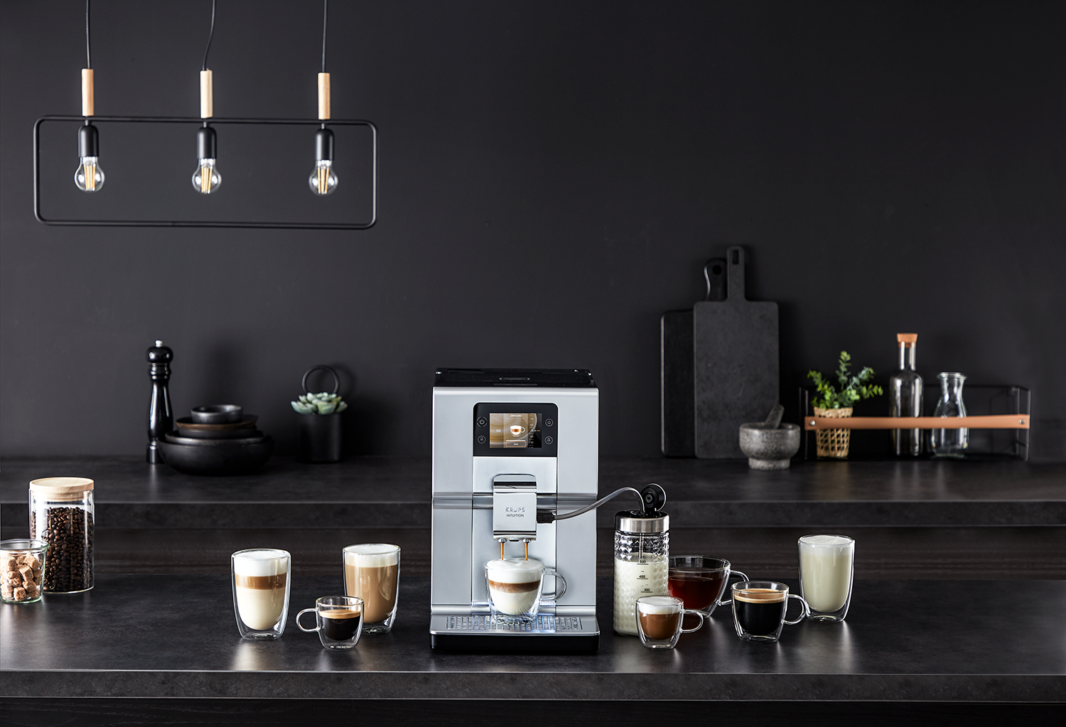 Una macchina da caffè argentata su un piano di lavoro scuro, circondata da tazze di caffè e una caraffa di latte su sfondo nero.