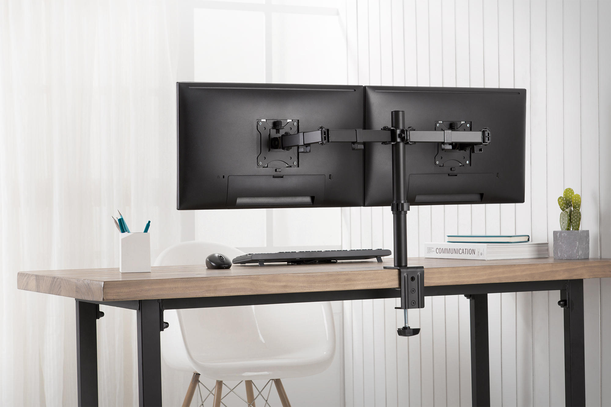Eine Doppelmonitorhalterung auf einem Holztisch mit Tastatur, Maus, Stiftehalter und einem kleinen Kaktus.