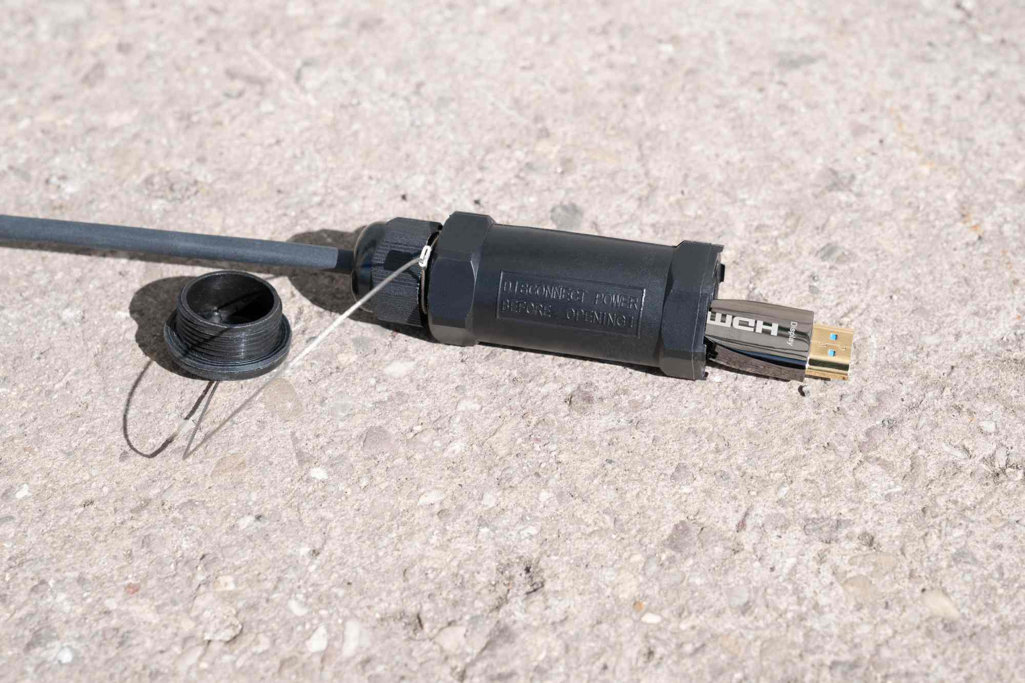 Ein schwarzes wasserdichtes HDMI-Kabelgehäuse mit abnehmbarer Kappe liegt auf hellem Beton.