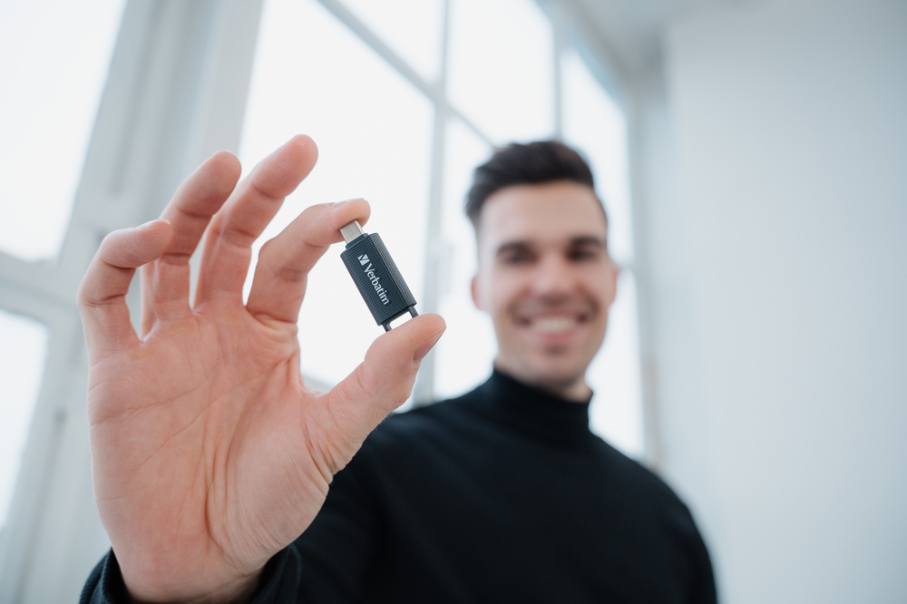 Un hombre sostiene una pequeña unidad USB negra en su mano, con una ventana brillante al fondo.