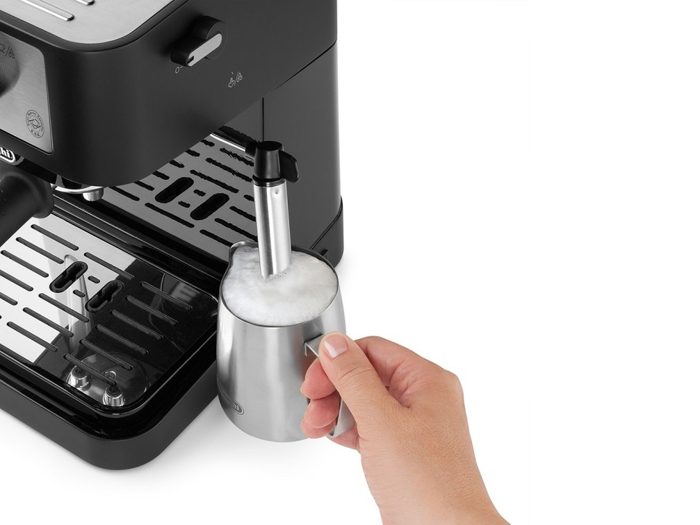 Una mano tiene una lattiera in acciaio inossidabile contro una macchina per espresso, montando il latte.