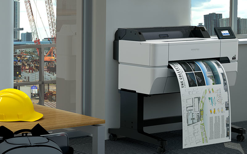 Una grande stampante plotter stampa un progetto in un ufficio con un cantiere visibile dalla finestra.