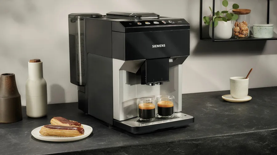 A black Siemens coffee machine stands on a kitchen counter with two espresso glasses and chocolate éclairs.