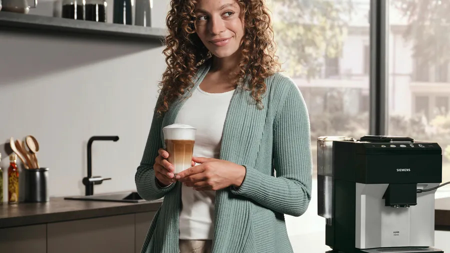 Curly-haired woman holding a latte macchiato in a kitchen with a coffee machine.
