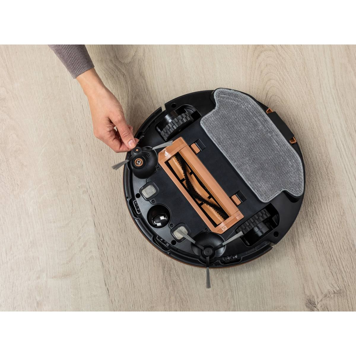 Mano retira un cepillo pequeño de la parte inferior de un robot aspirador negro en suelo de madera.