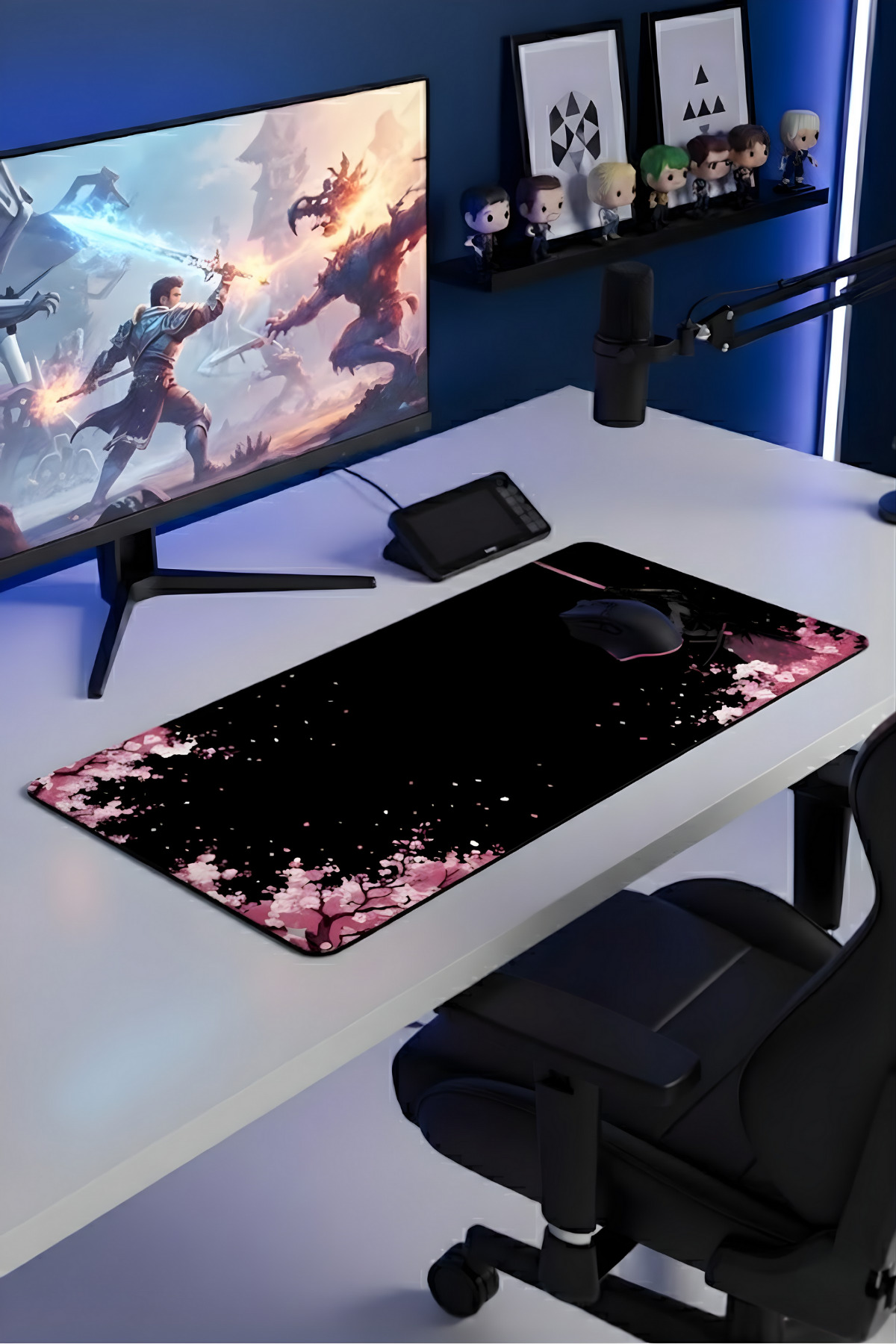 Fantezi sanatı gösteren monitörlü oyun masası, kiraz çiçekli siyah mousepad ve duvarda figürler.