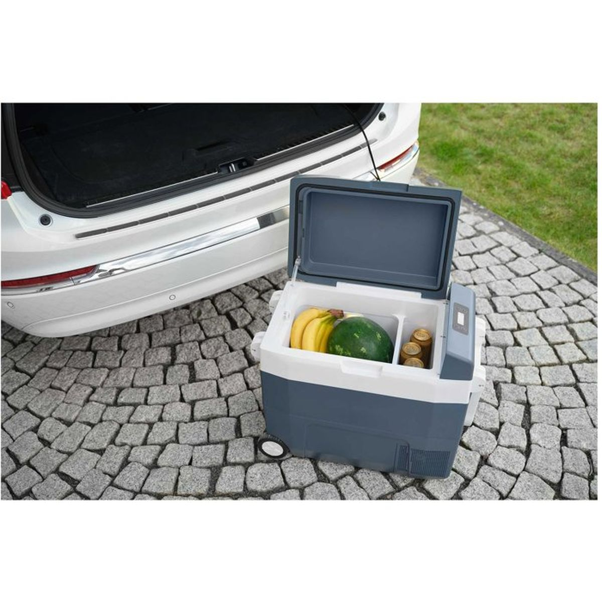 Eine graue Kühlbox mit Bananen, einer Melone und Dosen steht auf Pflastersteinen vor einem Auto.