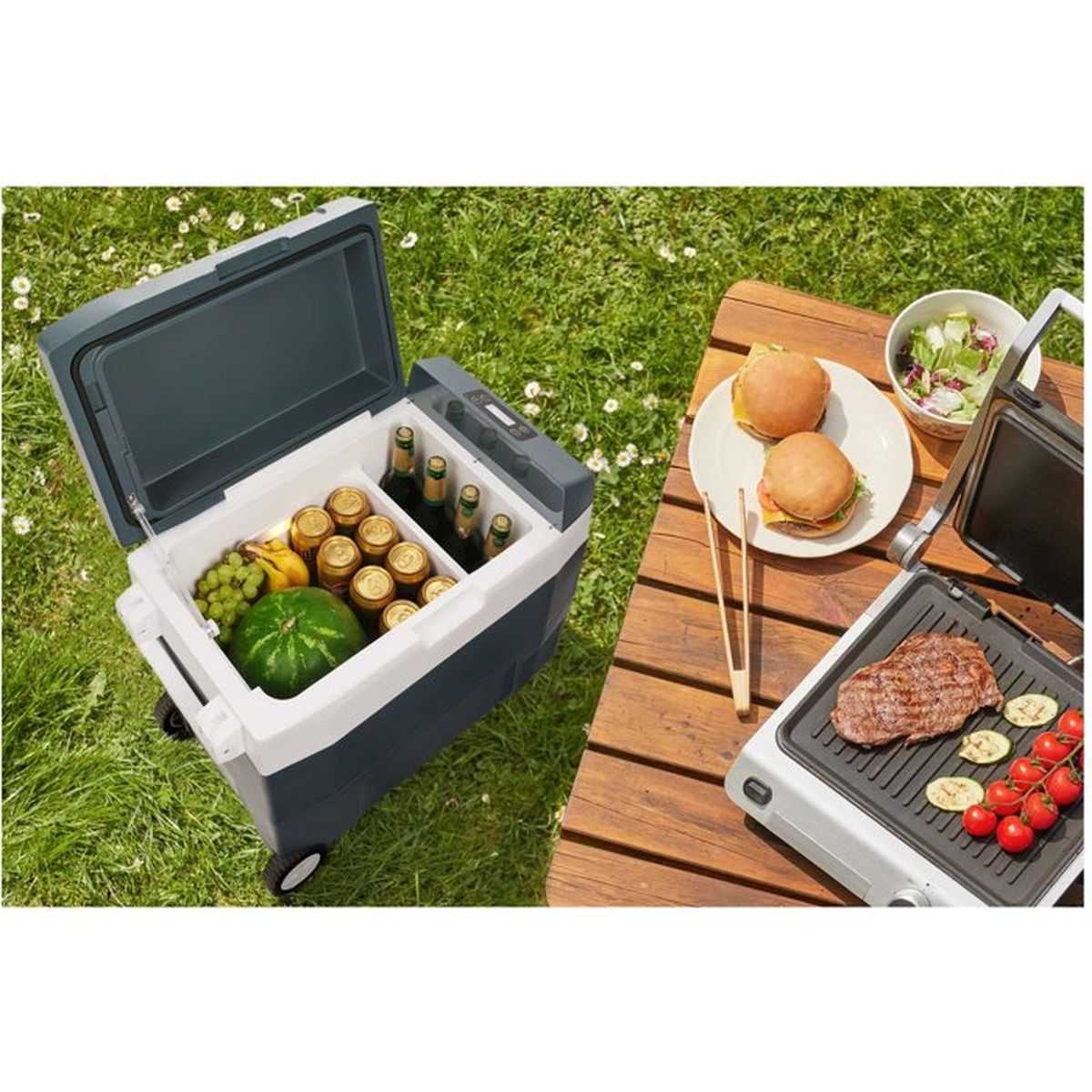 Eine offene Kühlbox mit Getränken und Obst steht auf Gras neben einem Holztisch mit Burger, Salat und Grill.