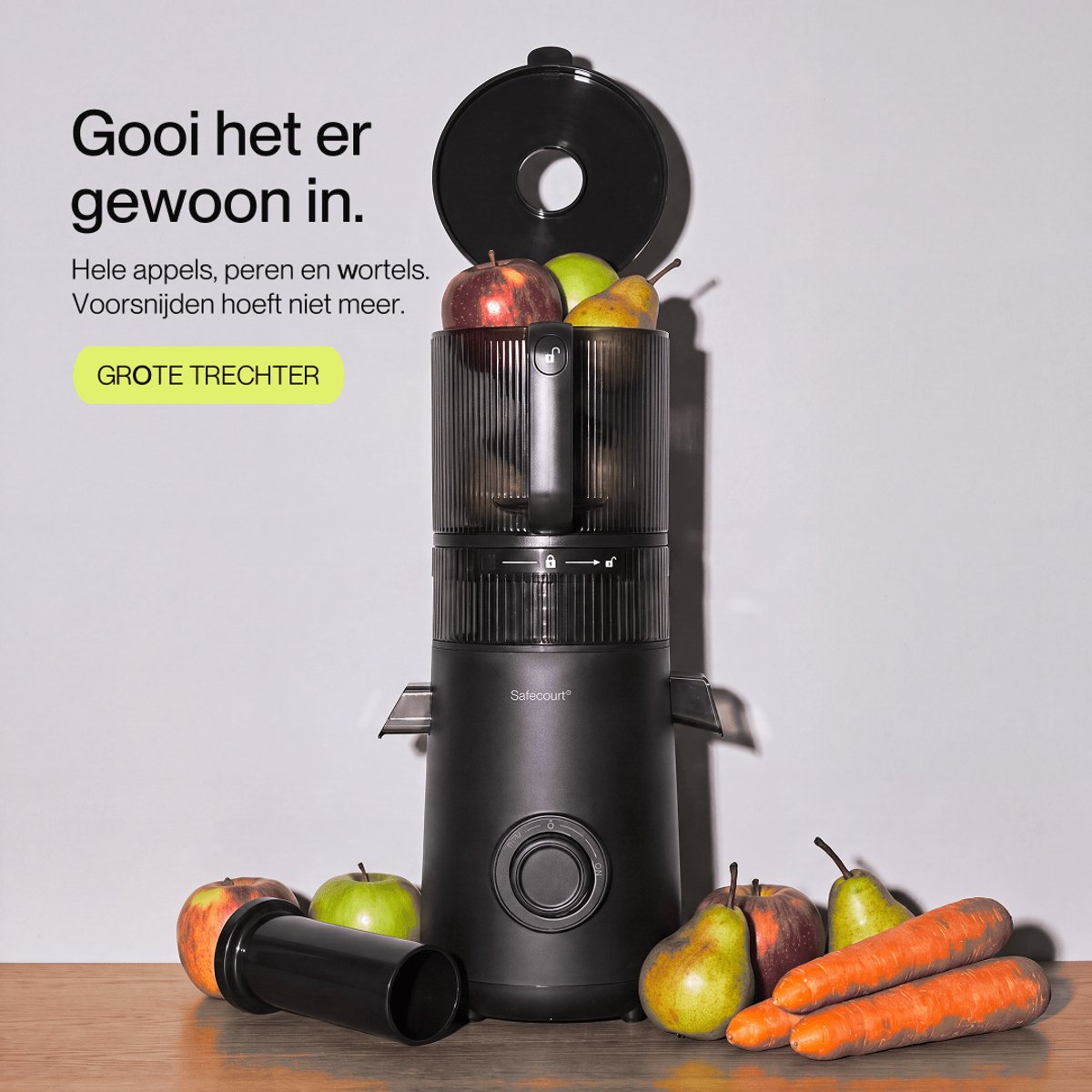 A black juicer with a large hopper is filled with apples and pears, with more fruit and carrots on the table.
