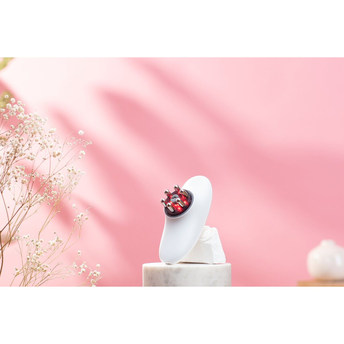 Ein weißes Hautpflegegerät mit Metallnoppen auf einem Marmorsockel vor rosa Hintergrund mit Trockenblumen.