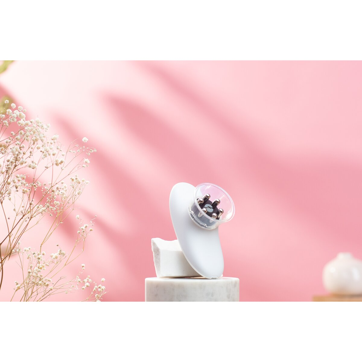 Ein weißes kosmetisches Gerät auf weißem Marmorsockel vor rosa Hintergrund mit Blumen.