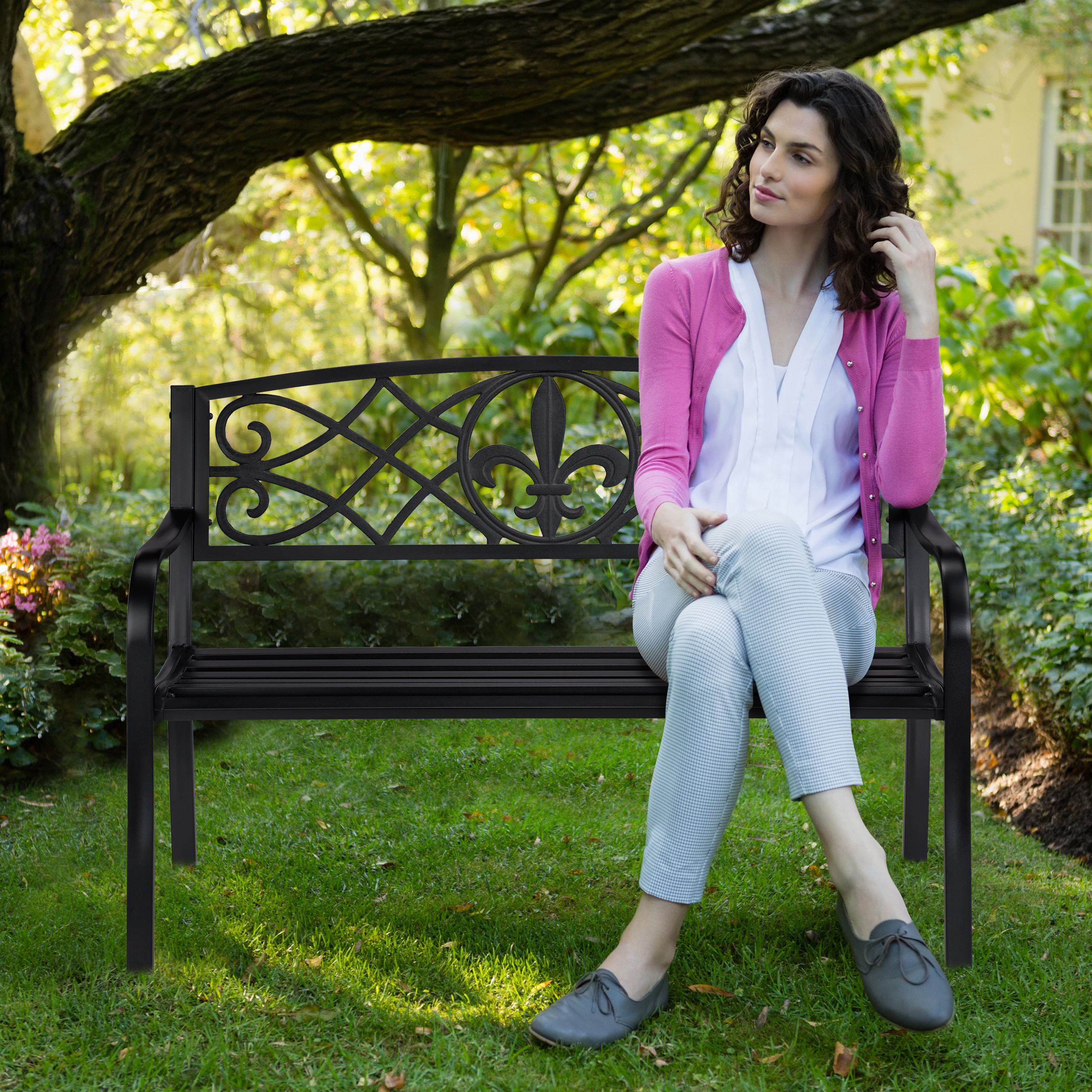 Eine Frau in rosa Strickjacke sitzt auf einer schwarzen Metallbank im Garten unter einem großen Baum.